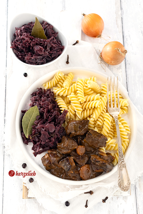 Weißer Teller mit einem deftigen Gulasch mit dunkler Soße nach einem Ofengulasch Rezept von herzelieb. Angerichtet mit Spiralnudeln und Rotkohl, der dekoriert ist mit Nelken und einem Lorbeerblatt.