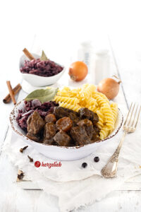 Eine Schale mit deftigem Rindergulasch mit dunkler Soße. Angerichtet mit Nudeln und Rotkohl nach einem Ofengulasch Rezept von herzelieb.