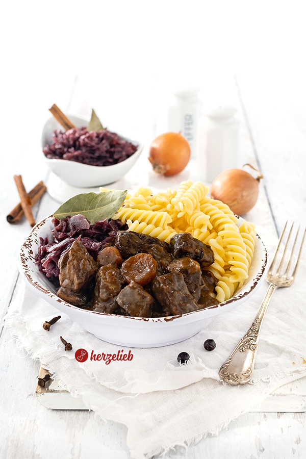 Eine Schale mit deftigem Rindergulasch mit dunkler Soße. Angerichtet mit Nudeln und Rotkohl nach einem Ofengulasch Rezept von herzelieb. 