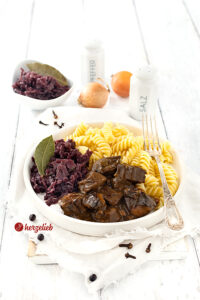 Ein Teller Gulasch mit dunkler Soße nach einem Ofengulasch Rezept von herzelieb. Angerichtet mit Nudeln und Rotkohl auf einem weißen Teller. Dekoriert mit einem Lorbeerblatt, ganzen Gewürznelken und Wacholderbeeren. Im Hintergrund eine Schale mit Rotkohl, zwei Zwiebel, einem Salzstreuer und einem Pfefferstreuer aus weiße,m Porzellan.