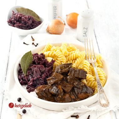 Ein Teller Gulasch mit dunkler Soße nach einem Ofengulasch Rezept von herzelieb. Angerichtet mit Nudeln und Rotkohl auf einem weißen Teller. Dekoriert mit einem Lorbeerblatt, ganzen Gewürznelken und Wacholderbeeren. Im Hintergrund eine Schale mit Rotkohl, zwei Zwiebel, einem Salzstreuer und einem Pfefferstreuer aus weiße,m Porzellan.