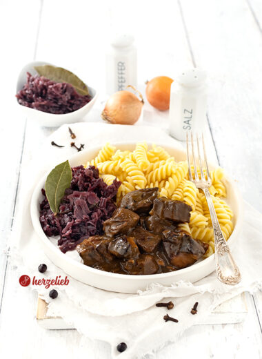 Ein Teller Gulasch mit dunkler Soße nach einem Ofengulasch Rezept von herzelieb. Angerichtet mit Nudeln und Rotkohl auf einem weißen Teller. Dekoriert mit einem Lorbeerblatt, ganzen Gewürznelken und Wacholderbeeren. Im Hintergrund eine Schale mit Rotkohl, zwei Zwiebel, einem Salzstreuer und einem Pfefferstreuer aus weiße,m Porzellan.