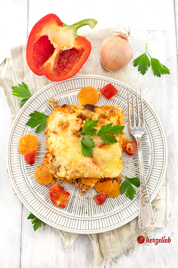 Auf diesem Bild zum Hackfleischauflauf Rezept sieht man den Reis-Hack-Auflauf auf einem Teller von oben fotografiert. Auf dem Tellerrand liegt ene Gabel. Aus dem Auflauf sind Karottenstücke, Paprikastücke und Hackfelisch geafllen. Im Hintergrund liegen eine halbe Paprikaschote und eine Zwiebel. 