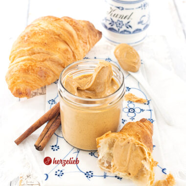 Glas mit hellbraunem Brotaufstrich nach einem Franzbrötchencreme Rezept von herzelieb. E steht auf auf einem Porzellanbrett mit weißblauem Muster, daneben ein Stück Croissant mit Creme. Im Hintergrund Zimtstangen, ein ganzes Croissant und ganz hinten ein Gewürzgefäß für Zimt mit weißblauem Dekor.