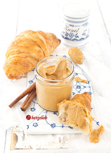 Glas mit hellbraunem Brotaufstrich nach einem Franzbrötchencreme Rezept von herzelieb. E steht auf auf einem Porzellanbrett mit weißblauem Muster, daneben ein Stück Croissant mit Creme. Im Hintergrund Zimtstangen, ein ganzes Croissant und ganz hinten ein Gewürzgefäß für Zimt mit weißblauem Dekor.