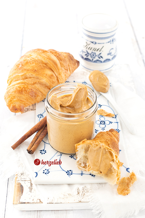 Glas mit hellbraunem Brotaufstrich nach einem Franzbrötchencreme Rezept von herzelieb. E steht auf auf einem Porzellanbrett mit weißblauem Muster, daneben ein Stück Croissant mit Creme. Im Hintergrund Zimtstangen, ein ganzes Croissant und ganz hinten ein Gewürzgefäß für Zimt mit weißblauem Dekor.