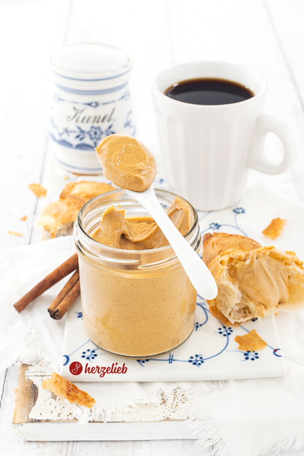 Glas mit Brotaufstrich nach dem Franzbrötchencreme Rezept von herzelieb.  Auf dem Glasrand liegt ein Löffel, neben dem Glas zerbröckelte Croissantstücke und eine Tasse schwarzer Kaffee, im Hintergrund Zimtstangen und ein blauweißes Gefäß für Zimt.