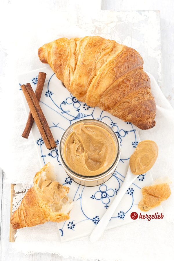 Hellbrauner Brotaufstrich im Glas nach einem Franzbrötchencreme Rezept von herzelieb. Franzbrötchen mit Creme, zwei Zimtstangen und ein Löffel. Vorne links ein halbes Croissant, mit dem Aufstrich bestrichen und im Hintergrund rechts ein ganzes. 