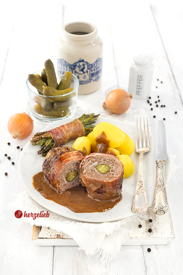 Eine halbierte Bauernroulade mit dunkler Soße nach einem Hackfleischrouladen Rezept von herzelieb.Die Roulade ist halbiert und auf einem Teller mit Speckbohnen und Kartoffeln und Soße angerichtet.