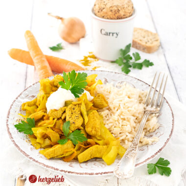 Auf einem Teller ist ein Kohlgericht mit Reis, zubereitet nach einem Karrykål Rezept aus Dänemark von herzelieb, angerichtet. Das Gericht enthält Hühnerstücke, durch Curry gelbfärbten Kohl und Karottenstücke. Oben drauf sind Sauerrahm und frische Kräuter. Serviert wird es mit Reis. Im Hintergrund steht ein Gewürzbehälter für Curry und auf dem Tellerrand liegt eine Gabel.