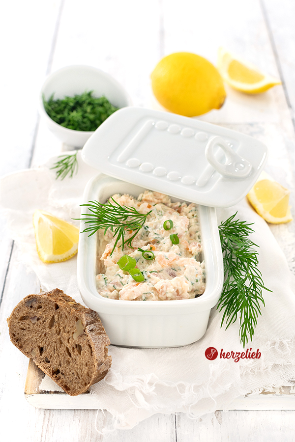 ein schneller Fischsalat nach einem Lachscreme Rezept von herzelieb. Angerichtet in einer rechteckigen, weißen Porzellandose mit Deckel. Dekoriert mit Dill , Zitronen und Brotscheiben 