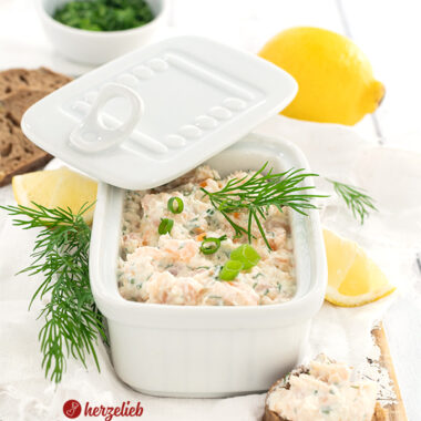 Fischsalat nach einem Lachscreme Rezept von herzelieb in einer eckigen, weißen Porzellanschale mit Deckel. Dekoriert mit Dill nund Zitronen. Im Vordergrund rechts ein bestrichees Baguettebrotstück.