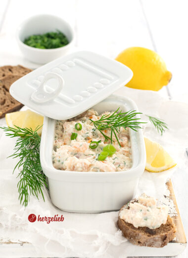 Fischsalat nach einem Lachscreme Rezept von herzelieb in einer eckigen, weißen Porzellanschale mit Deckel. Dekoriert mit Dill nund Zitronen. Im Vordergrund rechts ein bestrichees Baguettebrotstück.