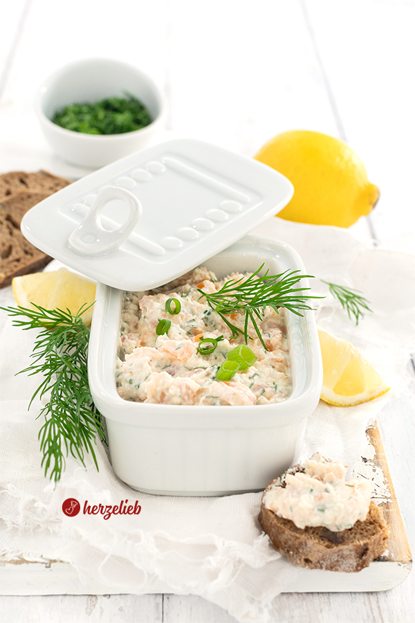 Fischsalat nach einem Lachscreme Rezept von herzelieb in einer eckigen, weißen Porzellanschale mit Deckel. Dekoriert mit Dill nund Zitronen. Im Vordergrund rechts ein bestrichees Baguettebrotstück.