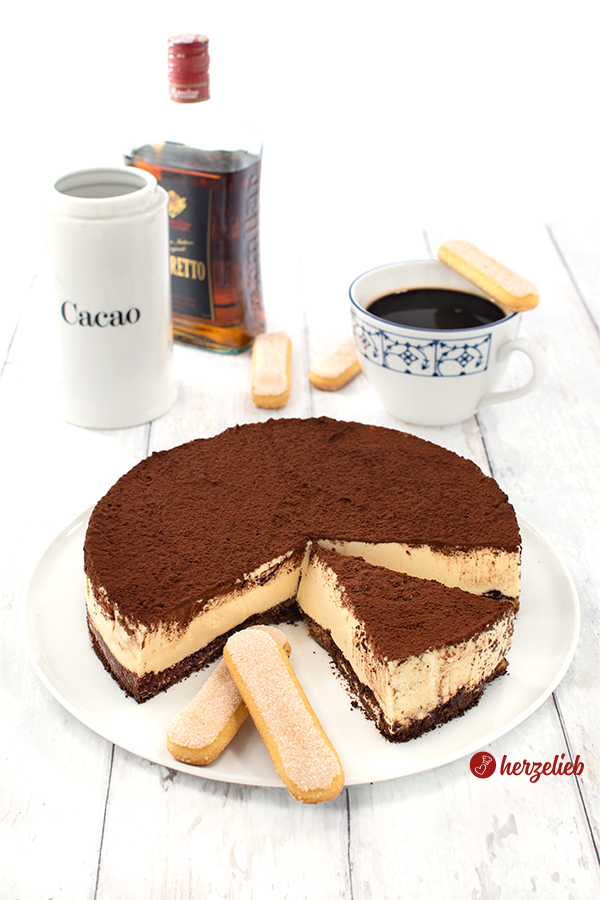 Eine angeschnittene Kühlschranktorte auf einer Tortenplatte nach einem Tiramisutorte Rezept von herzelieb.I Hintergrund eine Flasche Amaretto, ein Porzellangefäß mit Kakao. Daneben eine Tasse Kaffee.