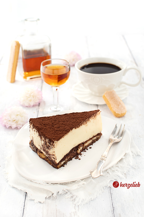 Ein Stück Kühlschranktorte nach einem Tiramisutorte Rezept auf einem weißen Teller. Auf dem Tellerrand liegt eine Kuchengabel. Im Hintergrund eine Tasse Kaffee, eine Flasche Amaretto, ein Likörgläschen sowie einige Blüten.