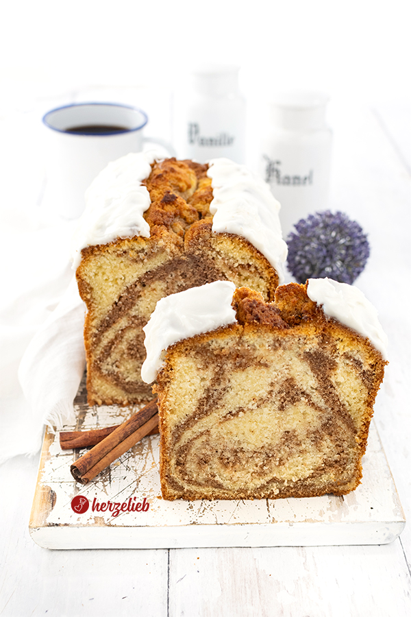 Angeschnittener Zimtkuchen nach einem Zimtschneckenkuchen Rezept. vor dem dem Kuchen steht ein einzelnes Stück aufrecht. Der Kuchen hat einen Schmandguss und steht auf einem weißen Holzbrett.