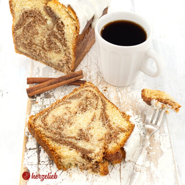 Im Hintergrund angeschnittener Rührkuchen nach einem Zimtschneckenkuchen Rezept. Im Vordergrund liegt ein Stück von dem Zimtkuchen, daneben eine Kuchengabel und dahinter steht eien Tasse Kaffee. Der Kuchen hat einen weißen Schmandzuckerguss