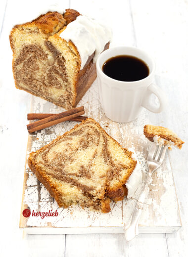 Im Hintergrund angeschnittener Rührkuchen nach einem Zimtschneckenkuchen Rezept. Im Vordergrund liegt ein Stück von dem Zimtkuchen, daneben eine Kuchengabel und dahinter steht eien Tasse Kaffee. Der Kuchen hat einen weißen Schmandzuckerguss