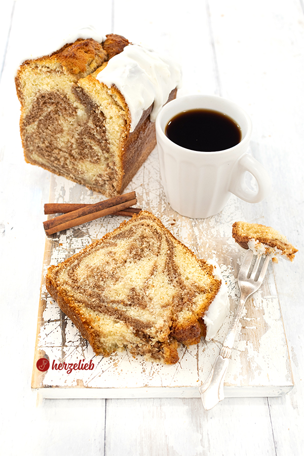 Im Hintergrund angeschnittener Rührkuchen nach einem Zimtschneckenkuchen Rezept. Im Vordergrund liegt ein Stück von dem Zimtkuchen, daneben eine Kuchengabel und dahinter steht eien Tasse Kaffee. Der Kuchen hat einen weißen Schmandzuckerguss