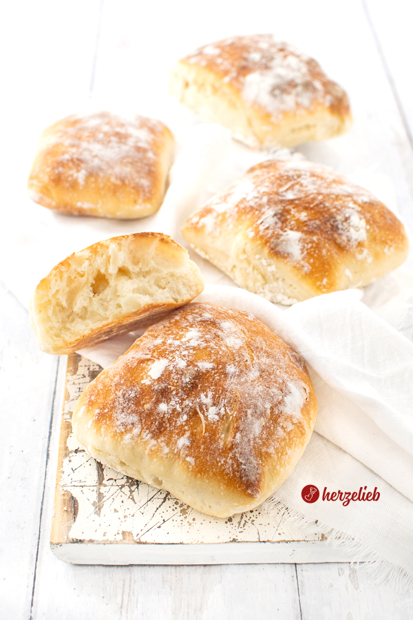 Vier Dinkel-Fladenbrötchen auf einem Holzbrett, das mit einem weißen Tuch belegt ist. Auf dem vorderen Brötchen liegt ein halbiertes Brötchen, bei dem man die Krume sieht.