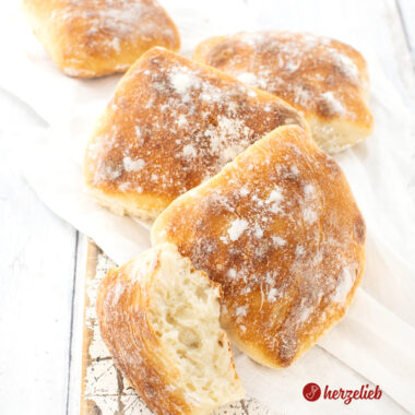 Knusprige Dinkel-Fladenbrötchen auf einem weißen Tuch, dass auf einem weißen Brett liegen. Vor den vier Brötchen liegt ein halbes Brötchen, bei dem man gut die Krume sieht.