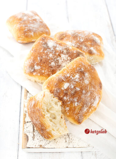 Knusprige Dinkel-Fladenbrötchen auf einem weißen Tuch, dass auf einem weißen Brett liegen. Vor den vier Brötchen liegt ein halbes Brötchen, bei dem man gut die Krume sieht.