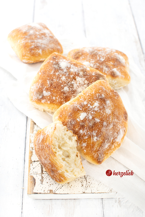 Knusprige Dinkel-Fladenbrötchen auf einem weißen Tuch, dass auf einem weißen Brett liegen. Vor den vier Brötchen liegt ein halbes Brötchen, bei dem man gut die Krume sieht.