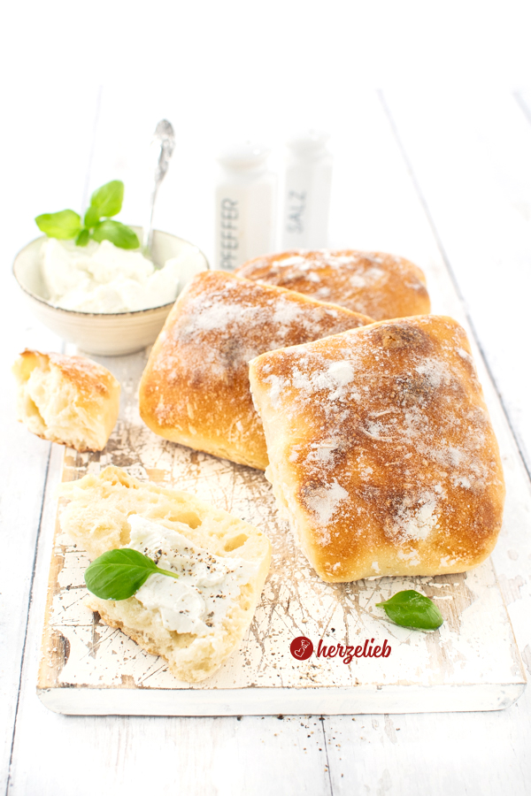 Drei frisch gebackenen Brötchen nach einem Dinkel-Fladenbrot Rezept von herzelieb auf einem Holzbrett. Links davor liegt ein mit Kräuterquark  bestrichenes Dinkelbrötchen. Im Hintergrund eine Schale mit Quarkaufstrich, ein Salz- und ein Pfefferstreuer.