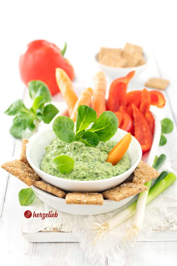 Eine weiße Schale mit einem grünen Dip nach einem Feldsalat-Dip von herzelieb. Angerichtet mit kleinen Möhren, Gemüsesticks und Crackern. Im Hintergrund eine schale mit Brotcrackern und eine ganze, rote Paprikaschote. Im Vordergrund rechts liegen zwei Lauchzwiebeln.Der Dip ist dekoriert Feldsalat