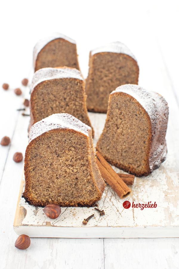 Fünf Stücke von einem Nusskuchen Gugelhupf nach einem Gewürz-Eilerikörkuchen Rezept von herzelieb. Die Gewürzkuchen Stücke  stehen aufrecht versetzt hintereinander auf einem weißen Holzbrett und sie sind mit Puderzucker bestäubt. Vor und neben den Stücken liegen Haelnüsse und Zimtstangen.