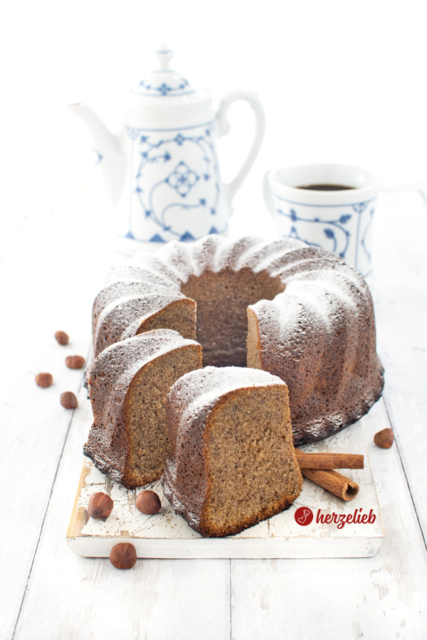 Ein Gugelhupf, der mit Puderzucker bestreut ist und nach einem Gewürz-Eierlikörkuchen Rezept von herzelieb gebacken wurde. Der Gewürzkuchen ist angeschnitten, zwei Scheiben stehen aufrecht halb davor.  Im Hintergrund steht weißblaues Kaffeegeschirr. Vor dem Nusskuchen liegen Haselnüsse und zwei Zimtstangen.