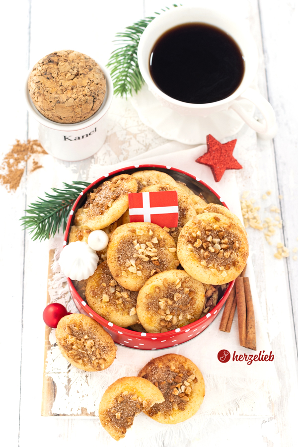 Eine Keksdose voll mit dänischen Weihnachtsplätzchen, die nach einem Jødekager Rezept aus Dänemark von herzelieb gebacken sind. Die Plätzchen in der Dose sind bestreut mit Zimt-Zucker und gehackten Mandeln, dazwischen steckt ein Dannebrog. Im Hintergrund eine Tasse Kaffee und ein Zimt-Gefäß. Vorne rechts zwei Zimtstangen und zwei weitere Weihnachtskekse.