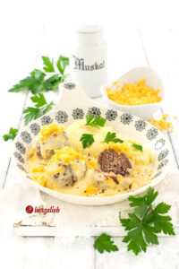 Dänische Hausmannskost nach einem Kødboller mit Ostesovs Rezept von herzelieb. Die Hackbällchen mit Käsesoße sind auf einem flachen Teller mit blauem Dekor angerichtet. Die Fleischbällchen liegen in einer gelben, cremigen Käsesoße vor einer Portion cremigen Kartoffelbrei, der mit Petersilie dekoriert ist. Im Hintergrund sieht man eine weiße Muskatnussdose aus Porzellan und eine kleine Schüssel mit geriebenem Käse.
