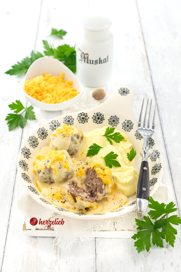 Hackbällchen in cremiger Käsesoße nach einem Kødboller mit Ostesovs Rezept von herzelieb. Angerichtet mit Kartoffelbrei, der mit Petersilie dekoriert auf einem flachen Teller mit blauem Dekor.  Im Hintergrund eine Muskatnussdose aus weißem Porzellan und eine Schüssel mit geriebenem Käse.