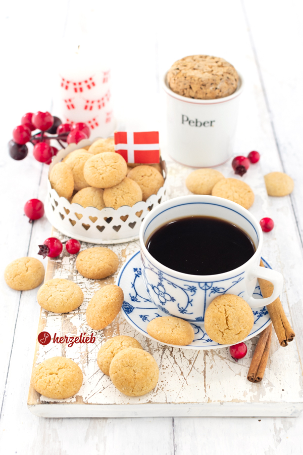 Eine Tasse Kaffee im Vordergrund, auf deren Unterteller zwei Pebernødder, gebacken nach einem Rezept aus Dänemark, liegen. Dahinter eine Schale mit dänischen Pfeffernüssen.