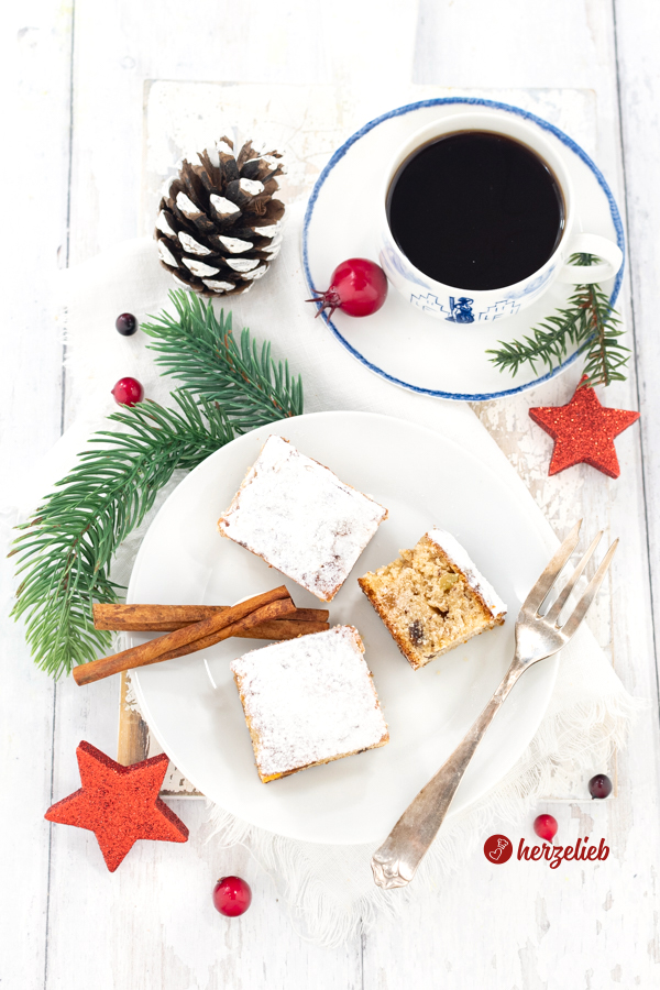 Drei Kuchenwürfel nach einem Stollenkuchen vom Blech Rezept auf einem weißen Teller. Sie sind von oben fotografiert und mit Puderzucker bestäubt. Auf dem Tellerrand liegt rechts eine Kuchengabel, links einige Zimtstangen. Dekoriert ist der Tisch mit Sternen, Tannnenzweigen und Tannenzapfen. Hinten rechts steht eine Tasse Kaffee.