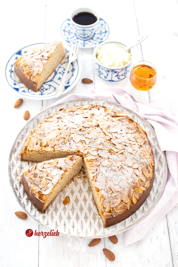 Ein angeschnittener Amarettokuchen auf einem weißen Teller mit grauem Muster. Im Hintergrund ein Teller mti einem Stück von dem Mandelkuchen, eine Tasse Kaffee und ein Gläschen Amaretto.