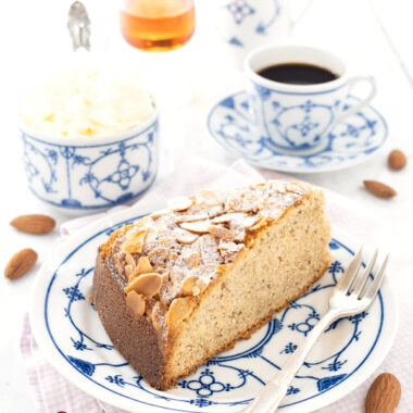 Ein Stück Mandelkuchen nach einem Amarettokuchen Rezept von herzelieb auf einem weißblauen Teller. Auf dem Tellerrand liegt eine Kuchengabel. Im Hintergrund eine Tasse Kaffee, eine Schale mit Schlagsahne, ein Likörglas und ein Milchkännchen