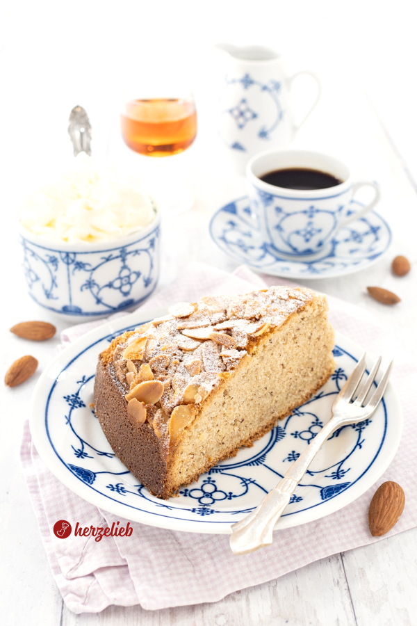 Ein Stück Mandelkuchen nach einem Amarettokuchen Rezept von herzelieb auf einem weißblauen Teller. Auf dem Tellerrand liegt eine Kuchengabel. Im Hintergrund eine Tasse Kaffee, eine Schale mit Schlagsahne, ein Likörglas und ein Milchkännchen