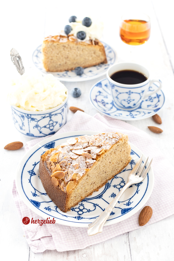 Zwei Teller mit jeweils einem Stück Amaretttokuchen. Dazwischen eine Tasse Kaffee, ein Schälchen mit Sahne. Das Stück im Hintergrund hat eine Sahnehaube und ist mit Heidelbeeren dekoriert.