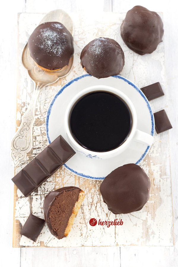 Dieses Bild zum Apelsinbiskvier Rezept aus Schweden von herzelieb ist von oben fotografiert. Rund um eien Tasse Kaffee sind Schokoladenkuchen, ein Schokoriegel und Schokoladenstücke verteil. Ein Kuchen ist halt durchgeschnitten und man sieht die Schichten aus Schokoladenüberzug, Schokocreme und einem Marzipanboden.