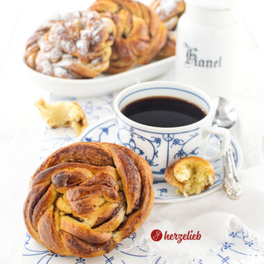 Ein goldbraun gebackener Kuchen, der wie eine Zimtschnecke ausschaut nach einem Karlsbaderbullar Rezept von herzelieb. Die Kuchenschnecke lehnt an einer Tasse Kaffee. Im Hintergrund weitere Kuchen auf einem länglichen Porzellanplatte.