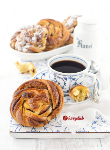 Ein goldbraun gebackener Kuchen, der wie eine Zimtschnecke ausschaut nach einem Karlsbaderbullar Rezept von herzelieb. Die Kuchenschnecke lehnt an einer Tasse Kaffee. Im Hintergrund weitere Kuchen auf einem länglichen Porzellanplatte.