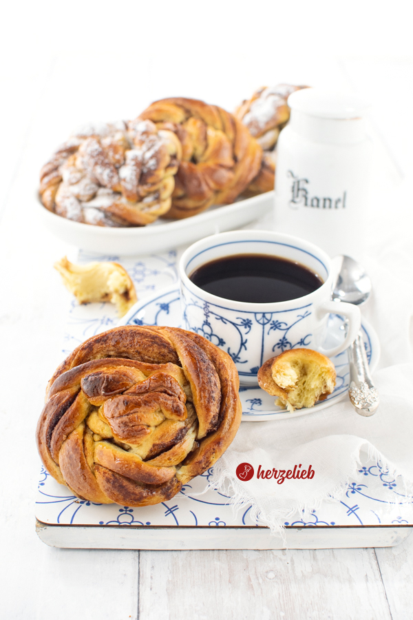 Ein goldbraun gebackener Kuchen, der wie eine Zimtschnecke ausschaut nach einem Karlsbaderbullar Rezept von herzelieb. Die Kuchenschnecke lehnt an einer Tasse Kaffee. Im Hintergrund weitere Kuchen auf einem länglichen Porzellanplatte.