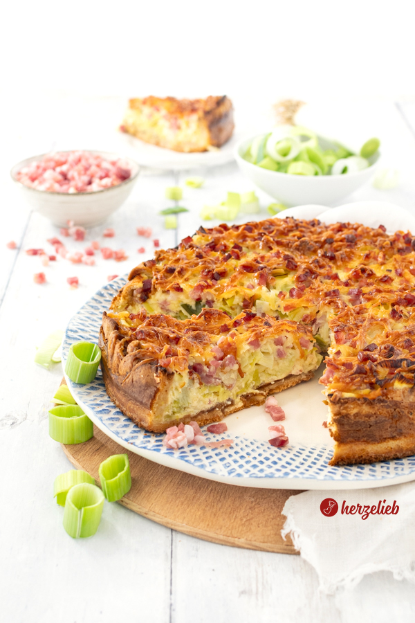 Angschnittene Lauchtarte mit Speck und Käse nach einem Porree-Tarte Rezept von herzelieb auf einem Teller. Ein Stück ist etwas nach vorne gezogen. Im Hintergrund eine Schale mit Schinkenwürfel und eine mit Lauchringen. Ganz hinten ein einzelnes Stück auf einem Teller.