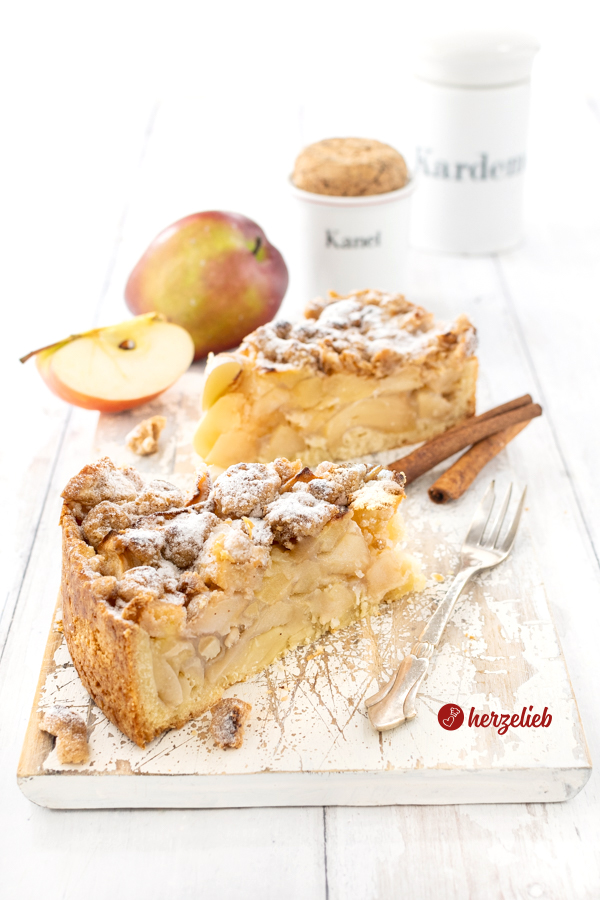 2 Stücke Apfelkuchen oder Streuselkuchen nach einem Apfel-Streuselkuchen Rezept von herzelieb. Im inneren sieht man die üppige Apfelfüllung, auf dem Kuchen die dicken Butterstreusel. Dekoriert Zimtstangen, Äpfeln und Gewürzgefäßen. Neben d den Kuchenstücken liegt eine Kuchengabel.