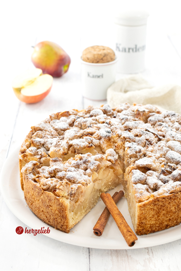 Ein angeschnittener Apfelkuchen nach einem Apfel-Streuselkuchen Rezept von herzelieb. Im Inneren ds Kuchens sieht man die üppige Apfelfüllung, obenauf die dicken Butterstreusel. Dekoriert mit Zimstangen. Im Hintergrnd ein ganzer Apfel und eine Apfelhälfte. Daneben zwei Gewürzgefäße aus Porzellan