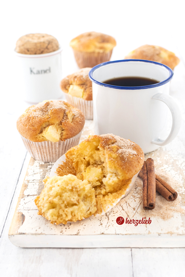 5 Apfelmuffins auf einem weißen Holzbrett nach einem Apfelkuchemuffins Rezept von herzelieb. Der vordere Muffin ist auseinandergebrochen, dahinter liegen zwei Zimtstangen. Weiter hinten ein weißer Becher mit blauem Rand mit Kaffee. Noch weiter hinten ein Gewürzgefäß mit einem Korken.