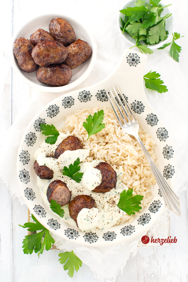 Ein Teller mit Hackbällchen mit Reis und Feta-Sahnesoße. Dekoriert mit glatter Petersilie. Auf dem Tellerrand liegt eine Gabel. Im Hintergrund eine Schale mit weiteren Fleischbällchen und ein Schälchen mit Peterslienblätten